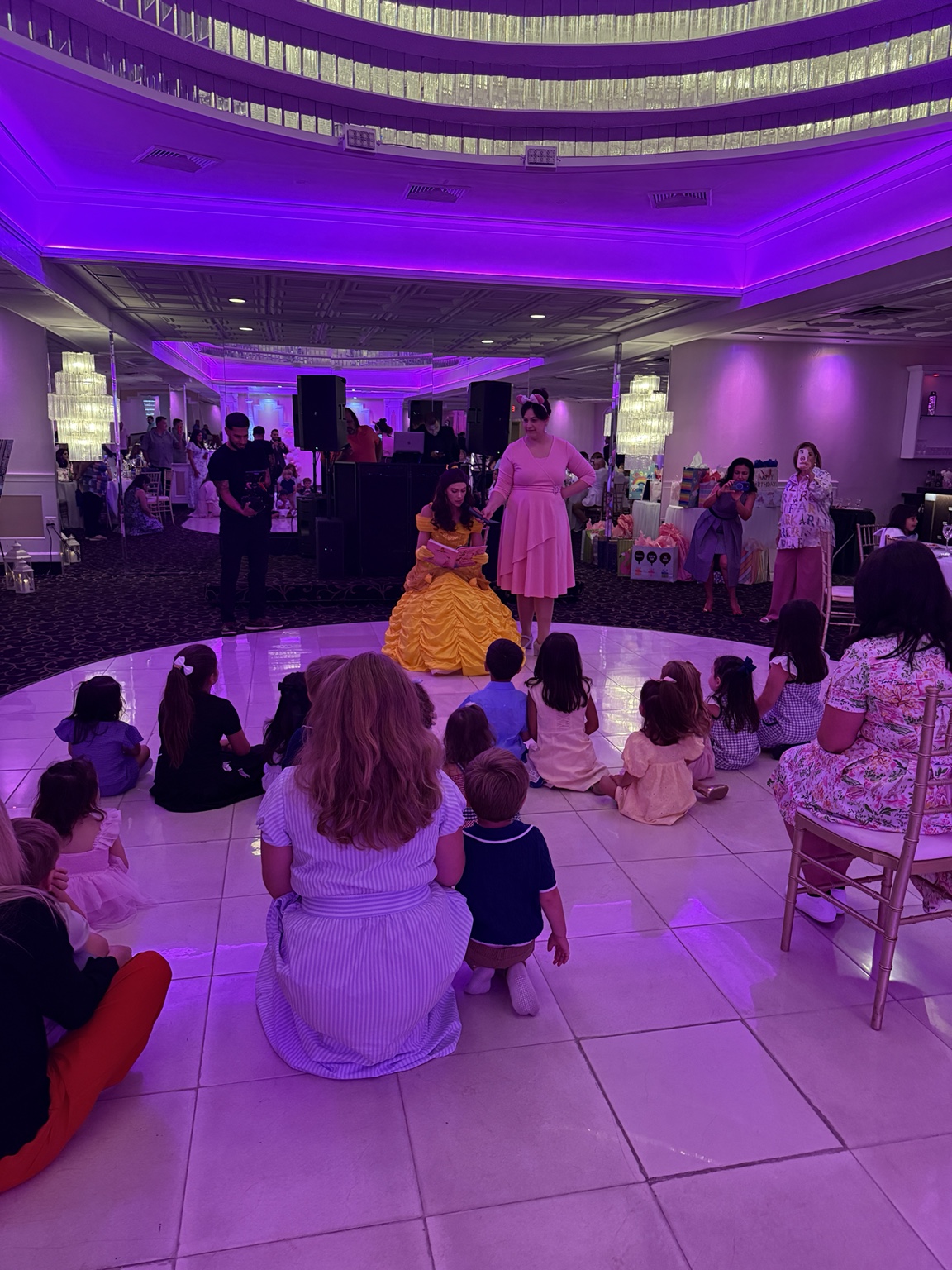 A princess in a yellow gown reading to a group of children at an indoor event