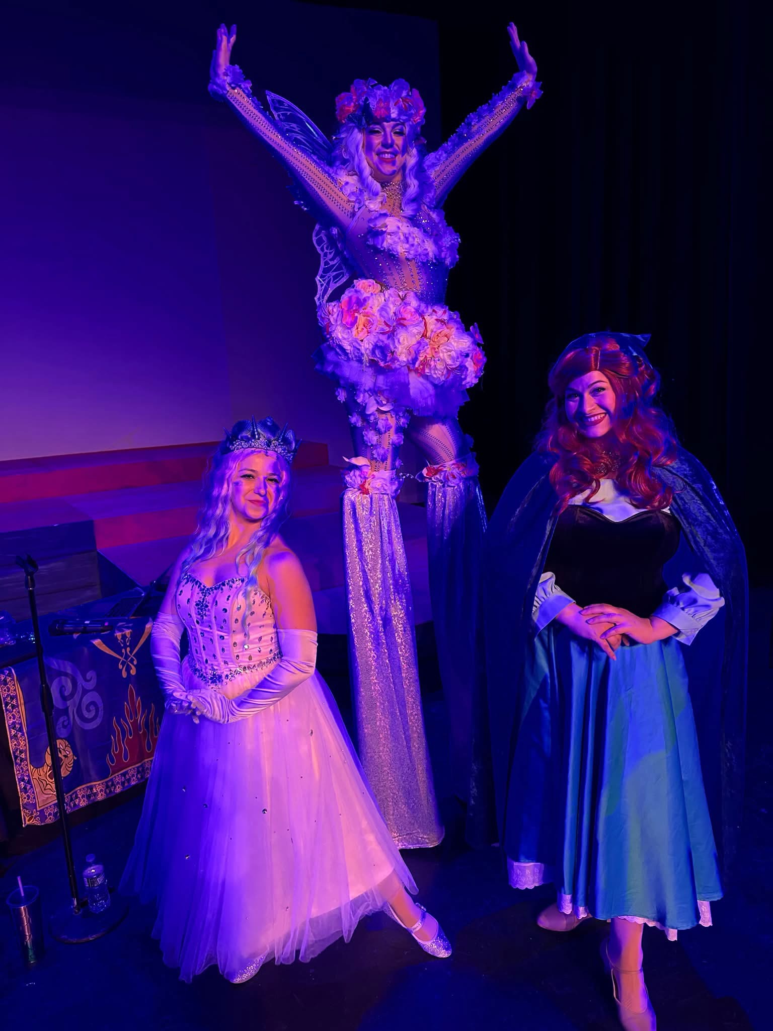 Three costumed performers under purple stage lights with a flower stilt walker