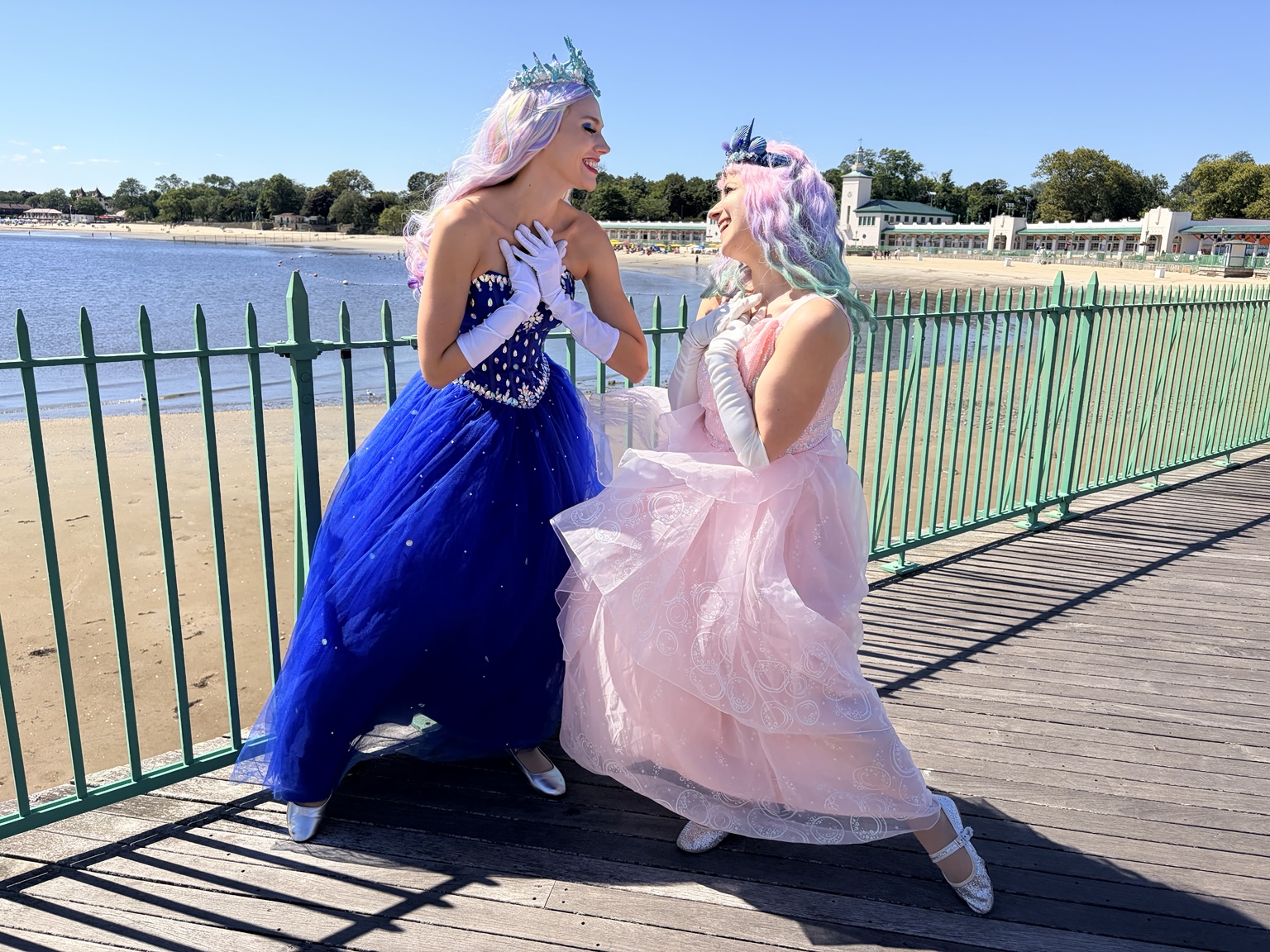 Two princess performers in blue and pink gowns smiling together by the waterfront